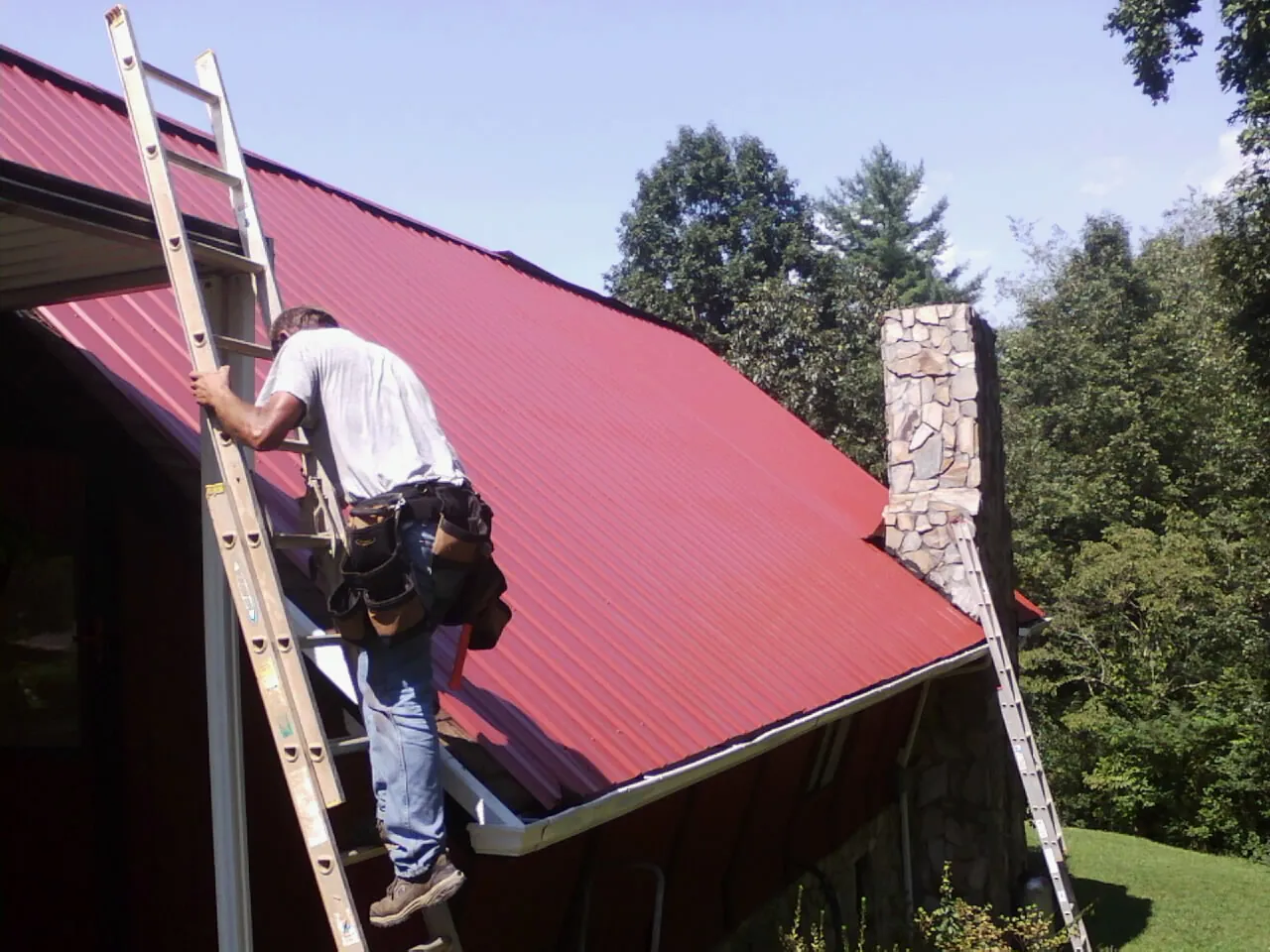Roofer on a ladder beside a red metal roof and stone chimney — Soffit & Fascia Repair by Roofing Brothers in Beech Grove